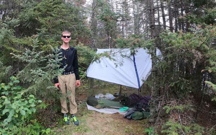 a student stands beside their tarp shelter in a wooded area on an outward bound trip